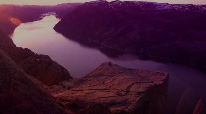 Two individuals standing on a cliff overlooking a fjord at sunset, immersed in meaningless conversations amidst the beautiful environment.